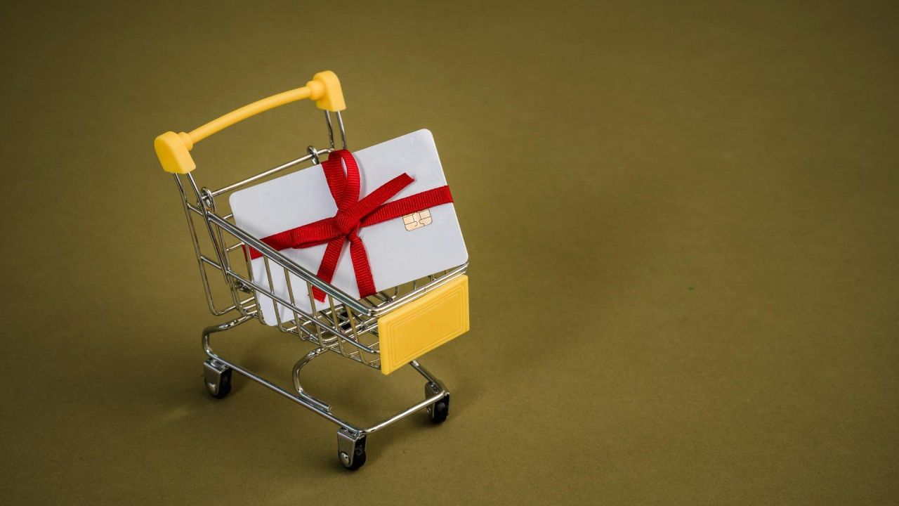 Gift card bianca con fiocco rosso dentro un mini carrello della spesa giallo, regalo digitale prepagato